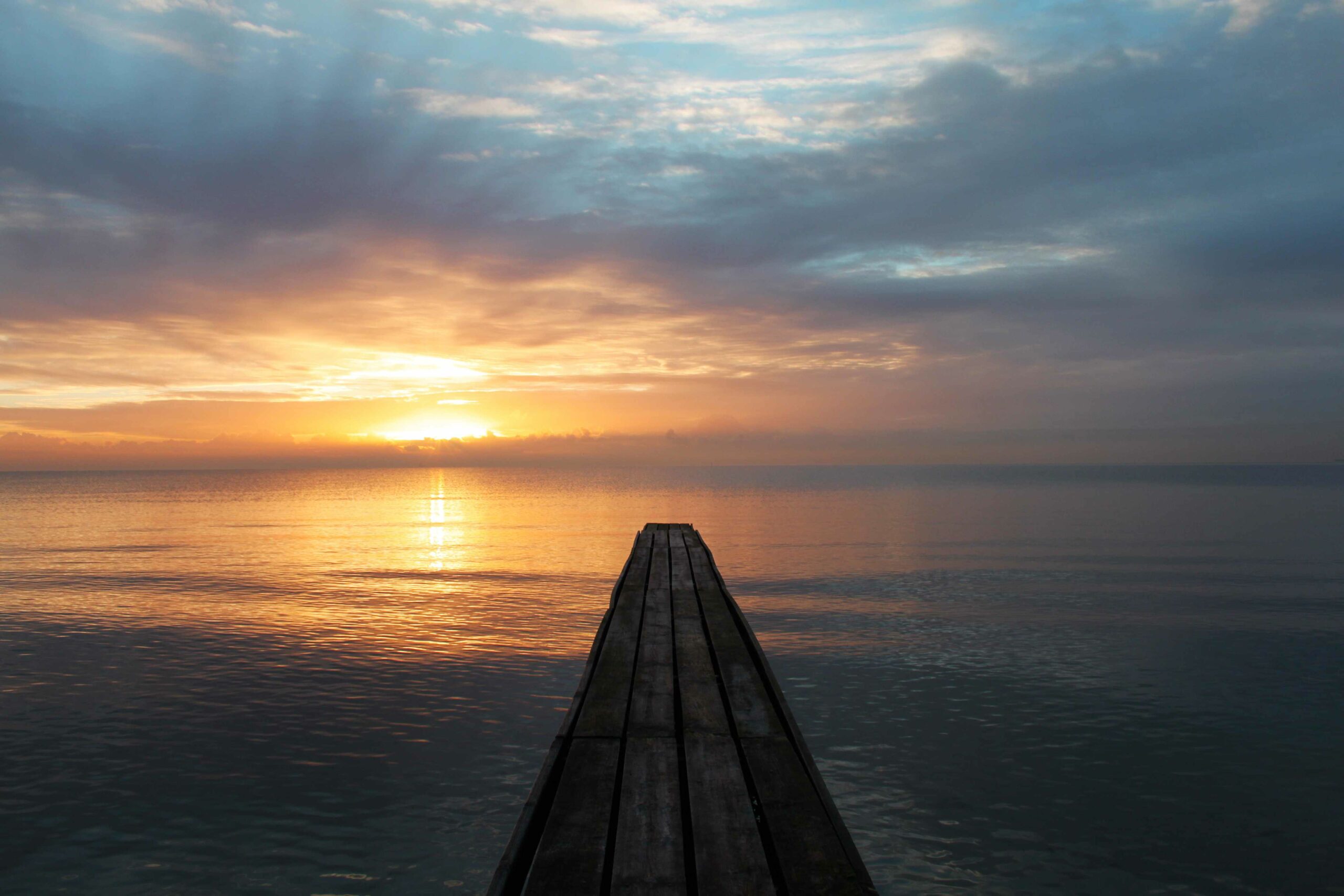 Das Bild zeigt einen Holzsteg bei Sonnenuntergang, welcher auf das Meer hinausgeht.